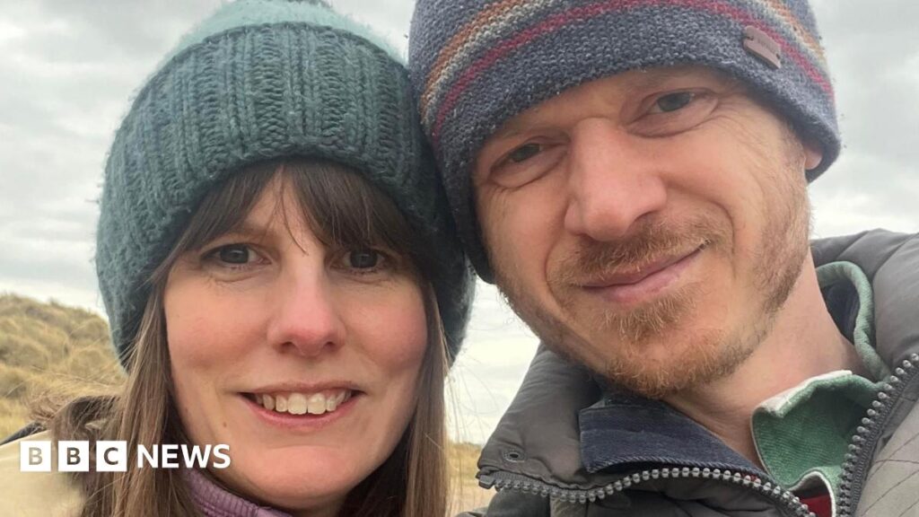 Annette Illing is standing next to her husband Mark on a beach on a cloud day. They are both wearing coats and knitted hats.