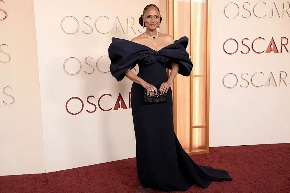 Person in an elegant off-the-shoulder gown poses on a red carpet at an award event backdrop