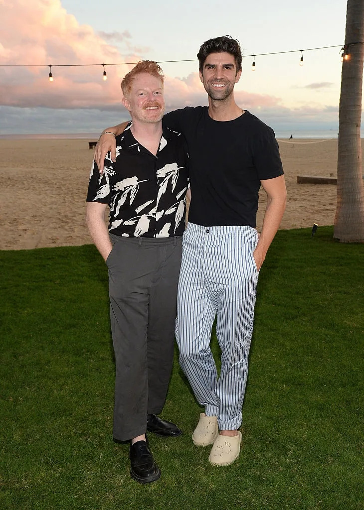 Two people smiling on grass by a beach at sunset. One wears a patterned shirt with pants, the other a solid shirt with striped pants