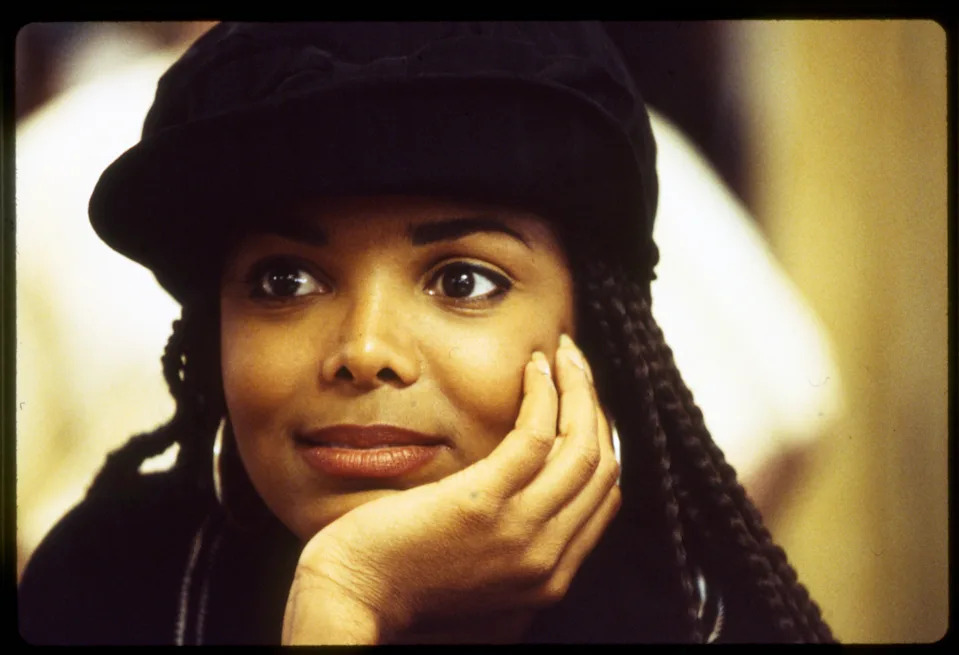 Janet Jackson with braided hair in a hat, resting chin on hand, gazing thoughtfully