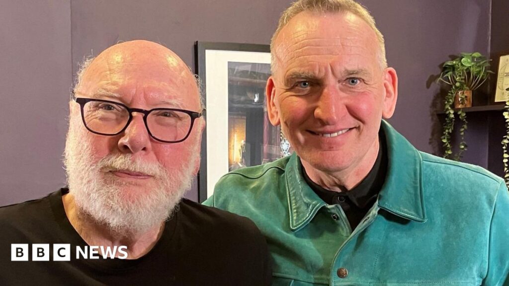 Peter Flannery and Christopher Eccleston standing side by side and smiling into the camera. Flannery is on the left and is bald with a white beard. He is wearing black-framed glasses and a black top. Eccleston is on the right and has grey hair and is wearing a green shirt. They are standing in front of a purple wall which has a photo frame hanging on it. There is a shelf with a plant in the right corner.