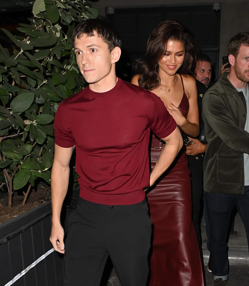 A man in a casual red shirt and black pants walks with a woman in a stylish red dress, possibly at a celebrity event