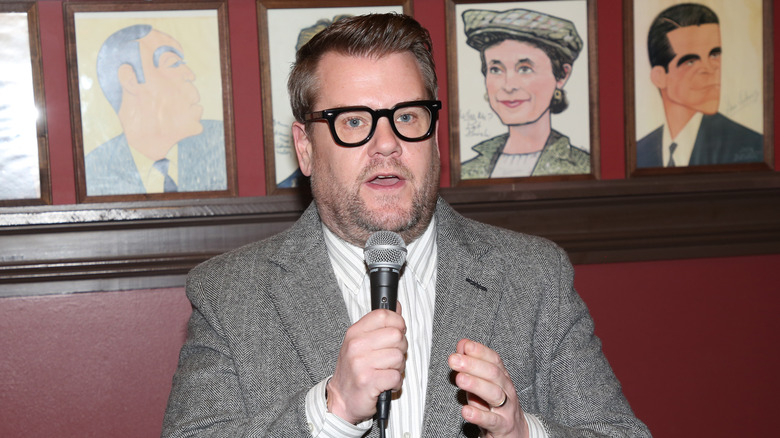 James Corden speaking in a mic and wearing glasses and a gray suit and striped shirt