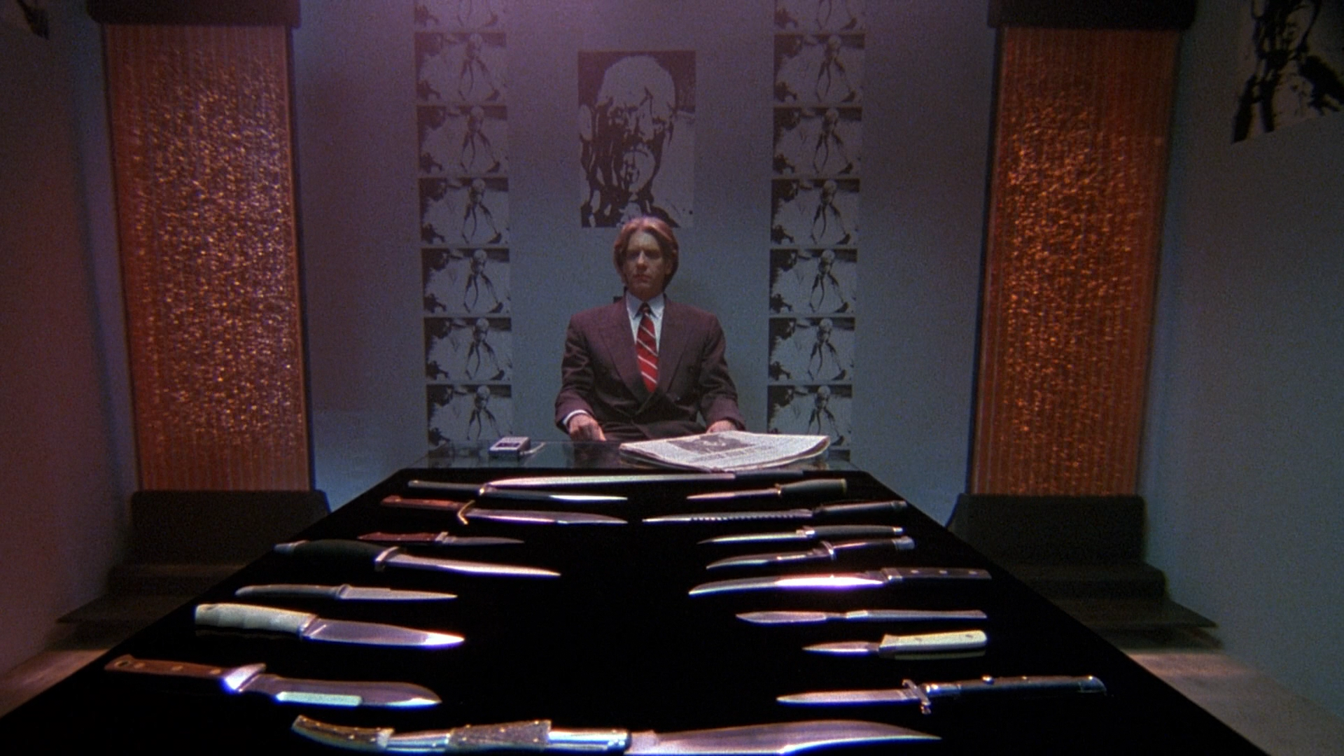 David Cronenberg looking at a table covered in blades, with two water sculptures flanking him and photos of grotesque art in the background.