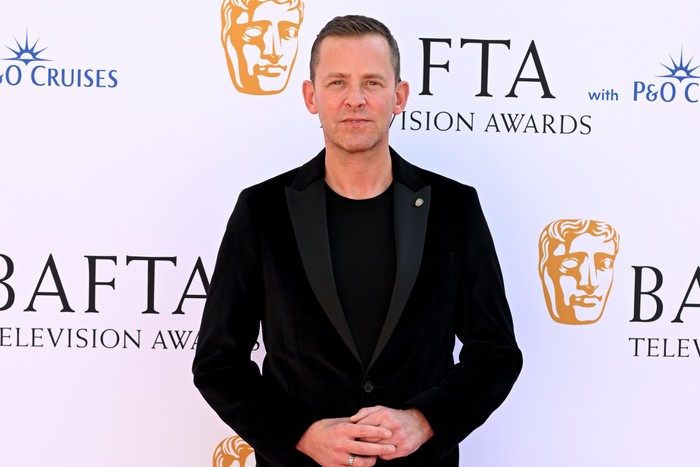 Scott Mills wearing an all-black tuxedo, smiling ahead with his hands placed together.