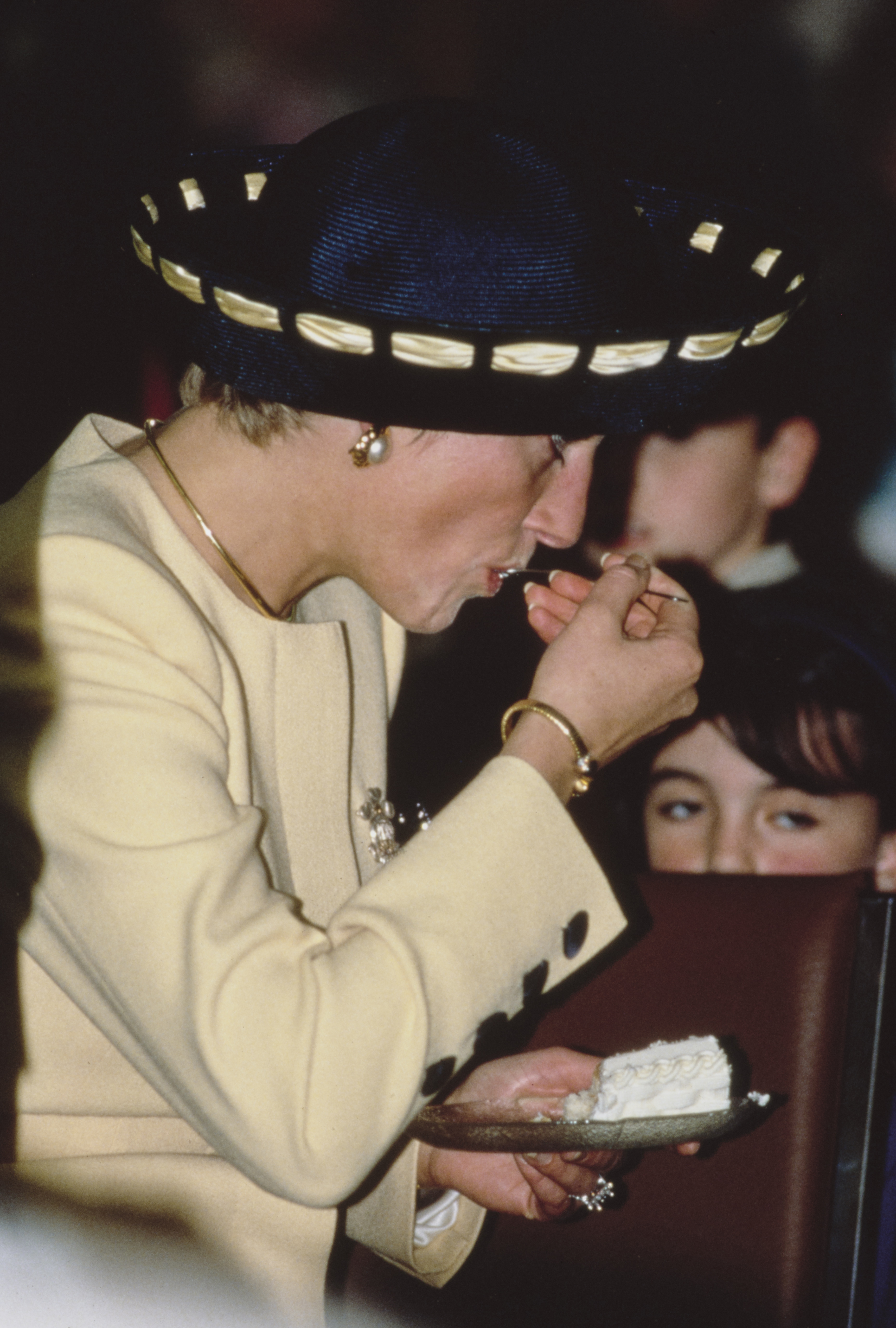 Princess Diana eating cake in a yellow coat and blue hat