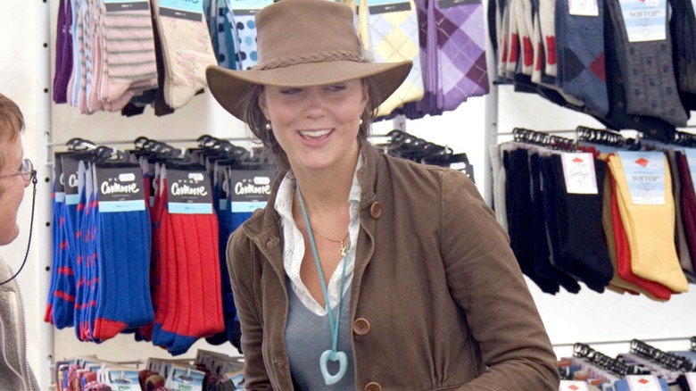 Kate Middleton wearing a brown hat and jacket over a shirt and sweater with a plastic heart necklace