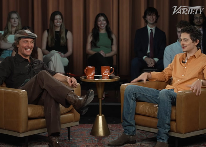 Steven Spielberg and Timothée Chalamet seated in armchairs during an interview with audience in the background.