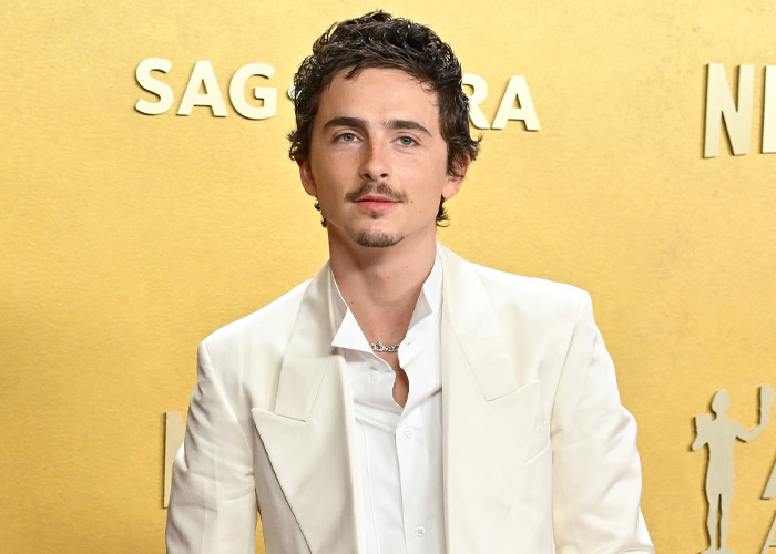 Timothée Chalamet in a white suit posing at the SAG Awards event against a gold background with SAG logo.