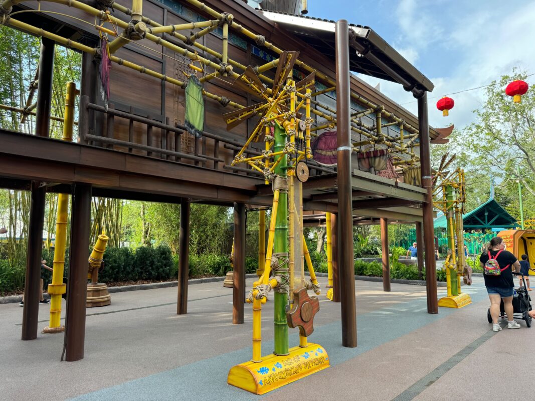 An outdoor structure with wooden beams and yellow mechanical installations underneath, surrounded by trees and a couple of people walking by. Red lanterns are hanging in the background, evoking the whimsical ambiance of DreamWorks Land.