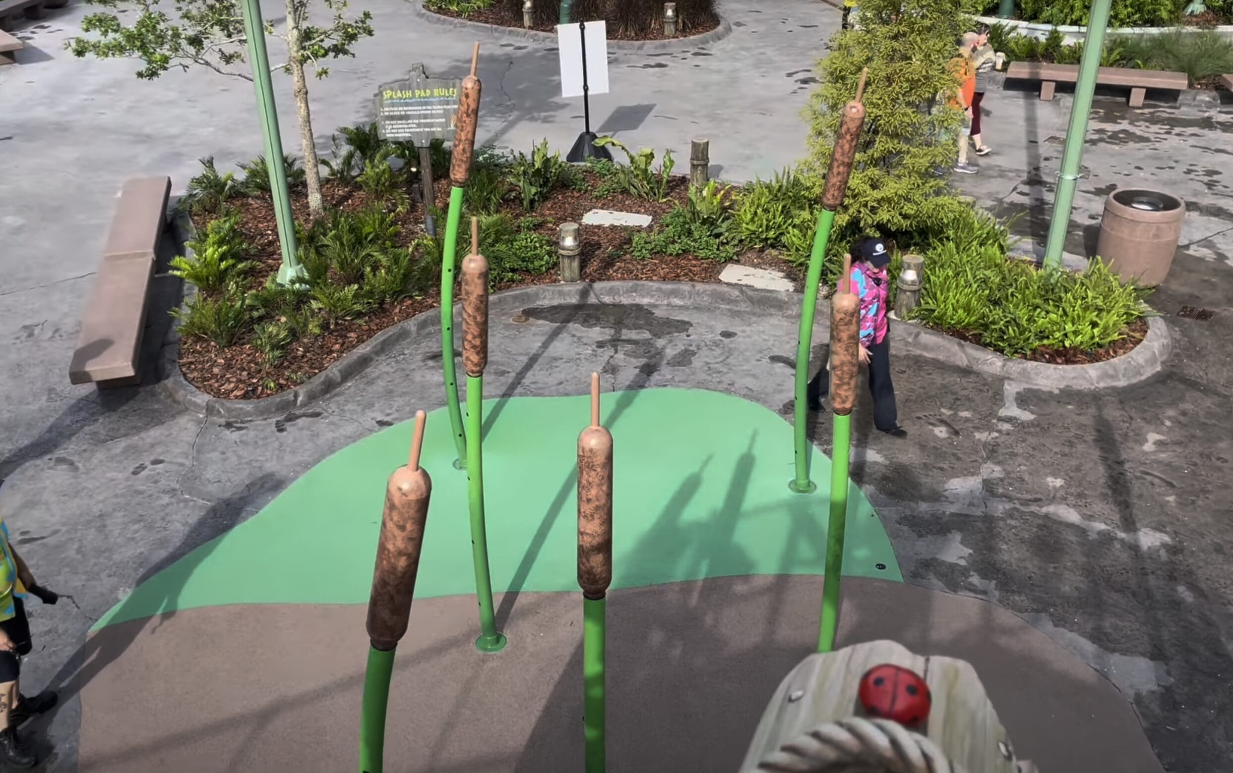 Large cattail sculptures on a green platform with people walking in a landscaped outdoor area.