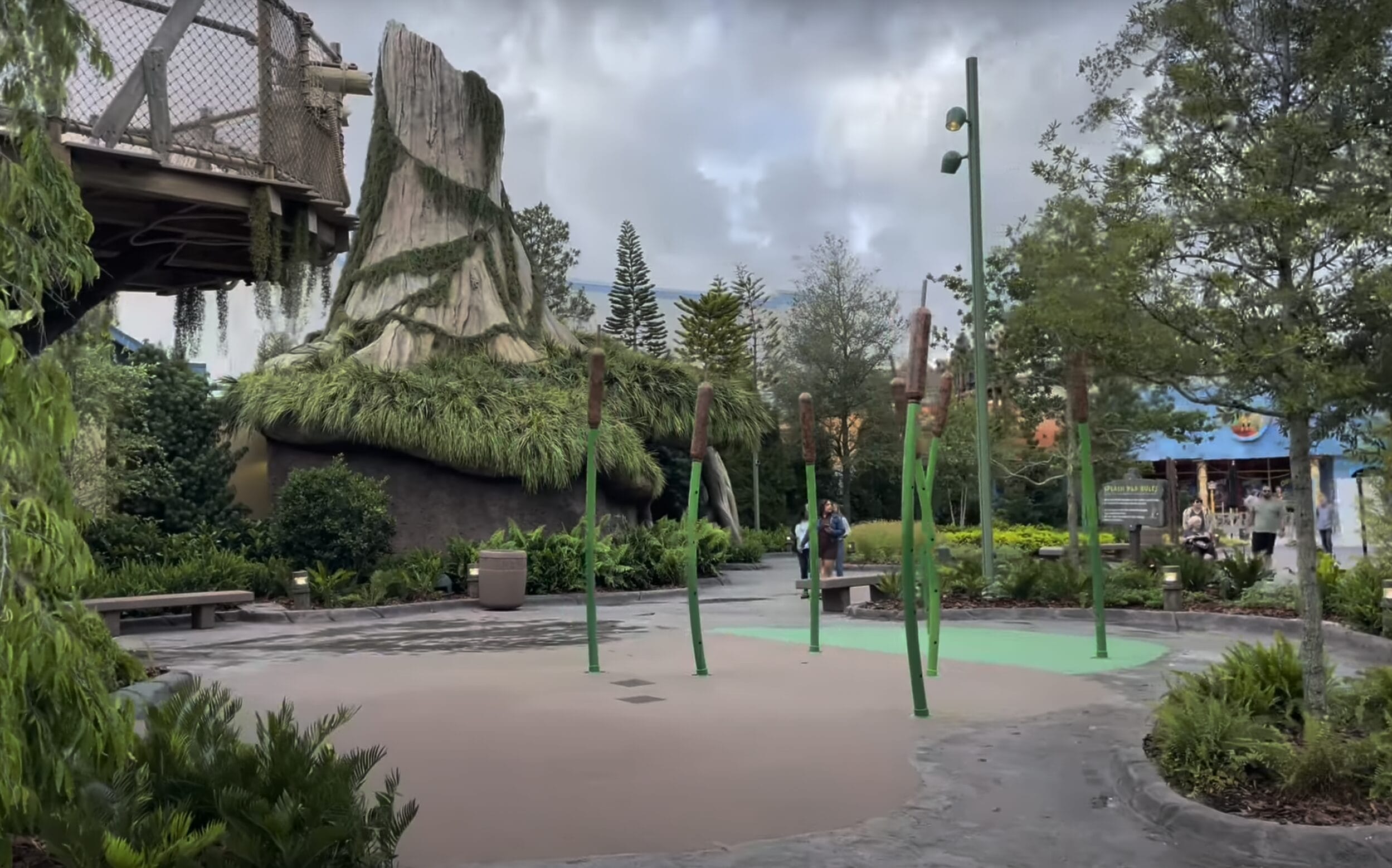 A themed outdoor splash pad with tall cattail-shaped fountains, greenery, rocks, and a cloudy sky.
