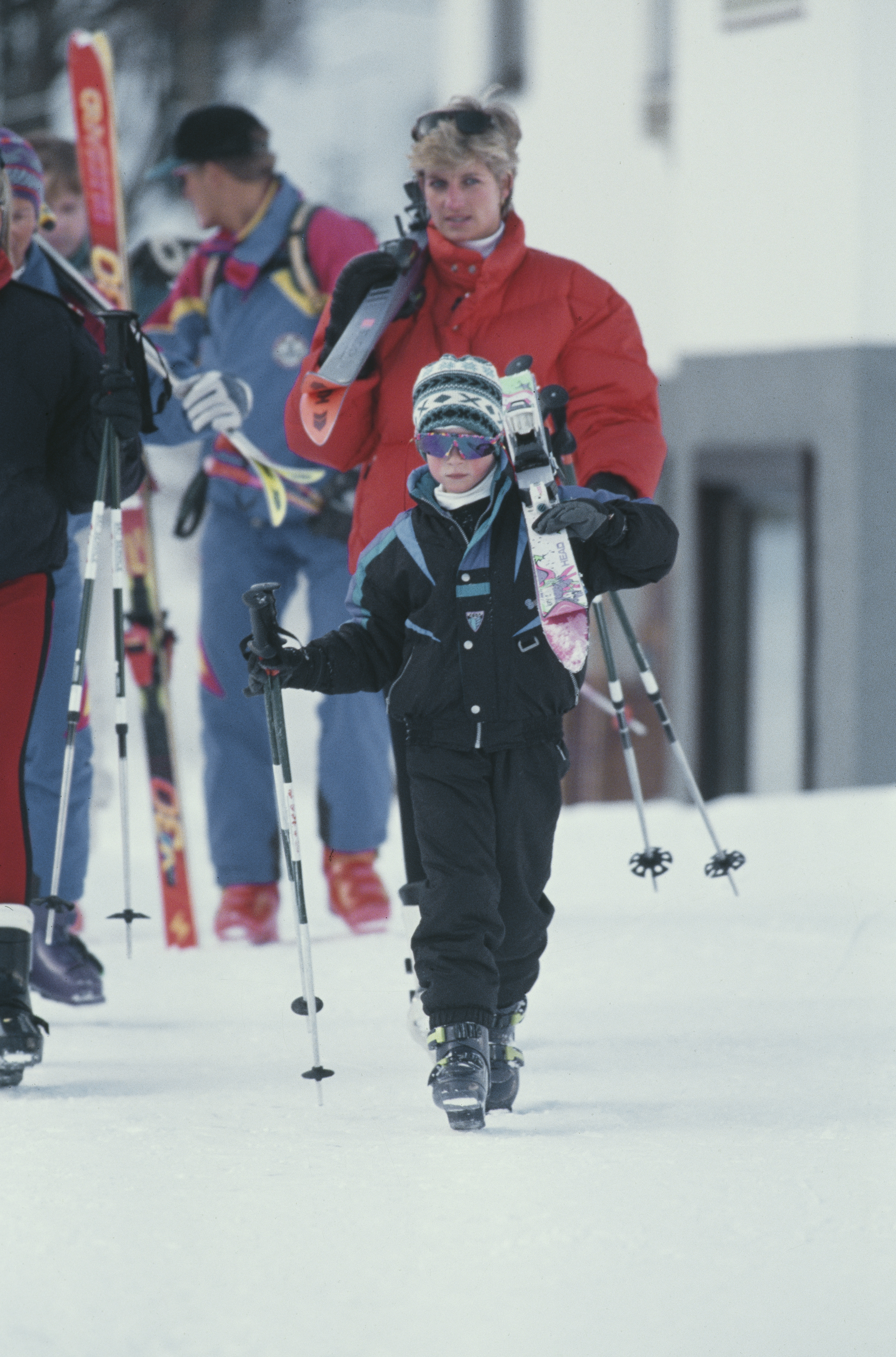 Prince Harry carrying skis with Princess Diana walking behind him