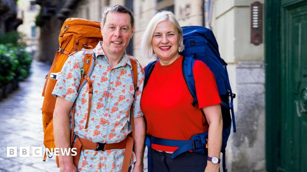 In-laws entered Race Across the World to ‘honour dying wish’ Mark Blythen, in his 60s, who has short grey hair and is wearing a short sleeved blue shirt with a lobster pattern and a large orange backpack, stands next to Margo Oakley, who has short blonde hair, a red t-shirt and a large blue backpack.