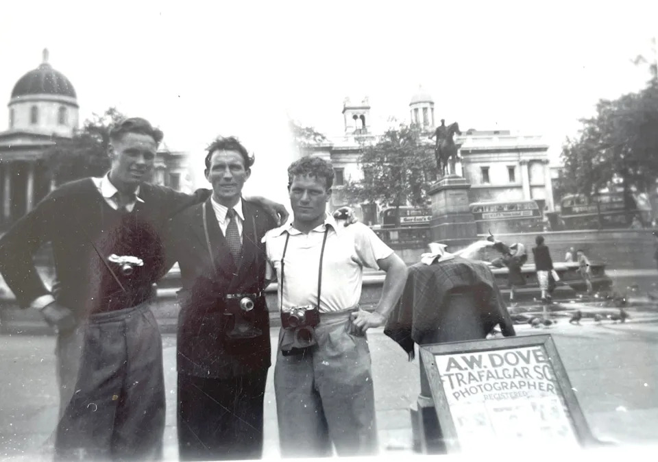 Three young men with cameras pose in front of the fountains at Trafalgar Square in 1950, with an A.W. Dove photographer's sign board propped beside them and the National Gallery visible in the background