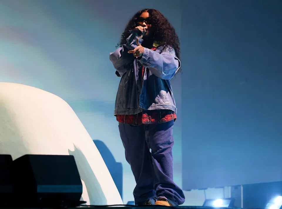 SZA performing on stage in oversized denim and plaid, holding a microphone with energetic expression