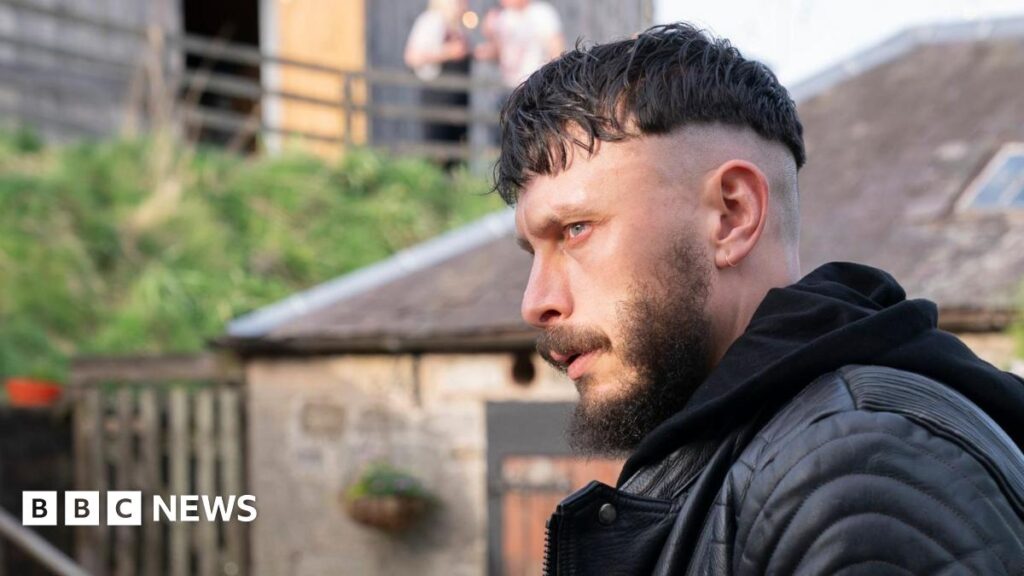 Richard Gadd stares intensely. He is wearing a black leather jacket and hoodie and is outside. Stone and slate buildings can be seen behind him. There are two people far behind on a terrace.