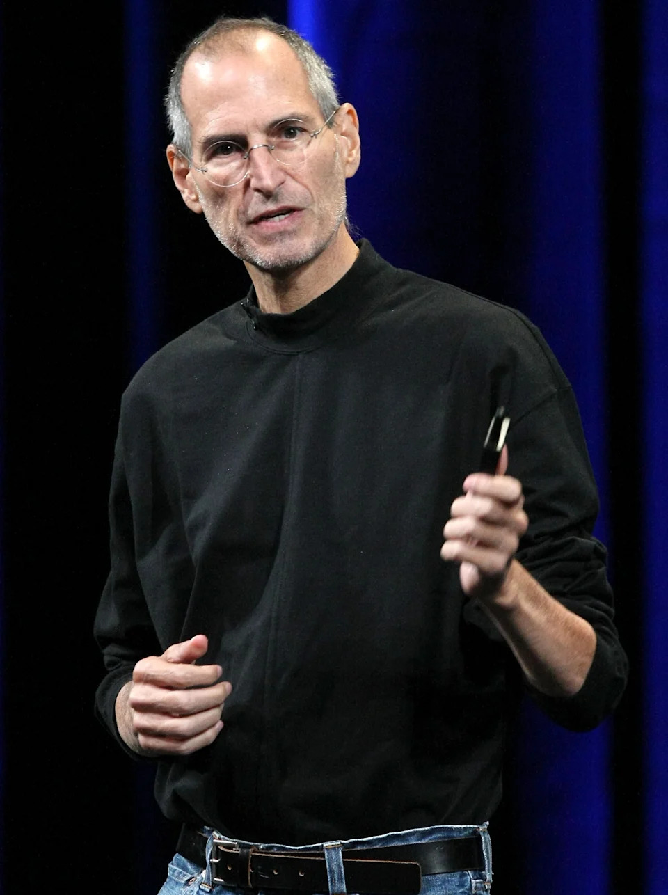A person in a black turtleneck and jeans holds a small object, speaking on stage at a presentation