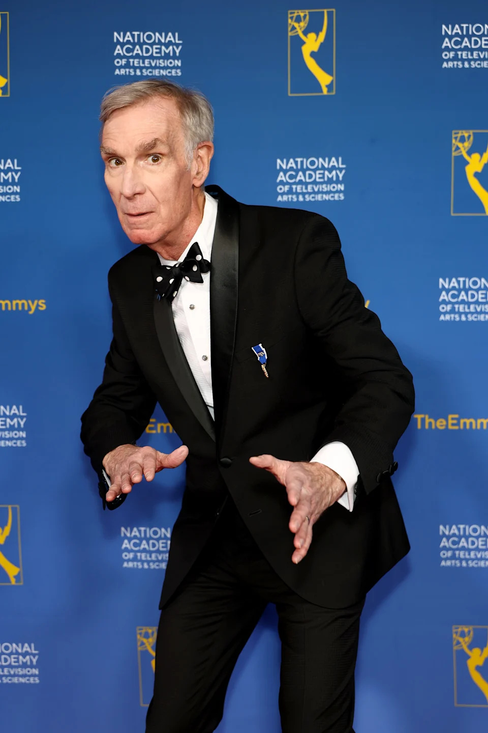 A man in a tuxedo with a bow tie poses playfully on the Emmys red carpet, set against a backdrop with Emmy logos