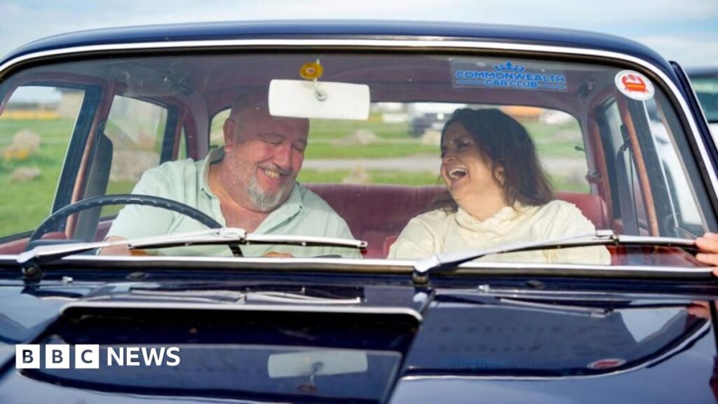 Jones and Speirs in an old navy vintage car laughing with each other. Jones, on the right, is in the passenger seat and is laughing whilst looking at Speirs. She wears a cream jumper and has brown hair. Speirs, is on the left, and wears a light green t-shirt and is looking down whilst laughing.