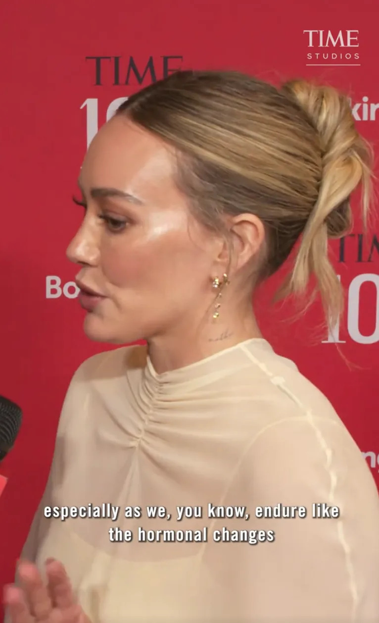 Person speaking during a red carpet event, wearing a sheer top with tied-back hair. Subtitles: "especially as we, you know, endure like the hormonal changes."