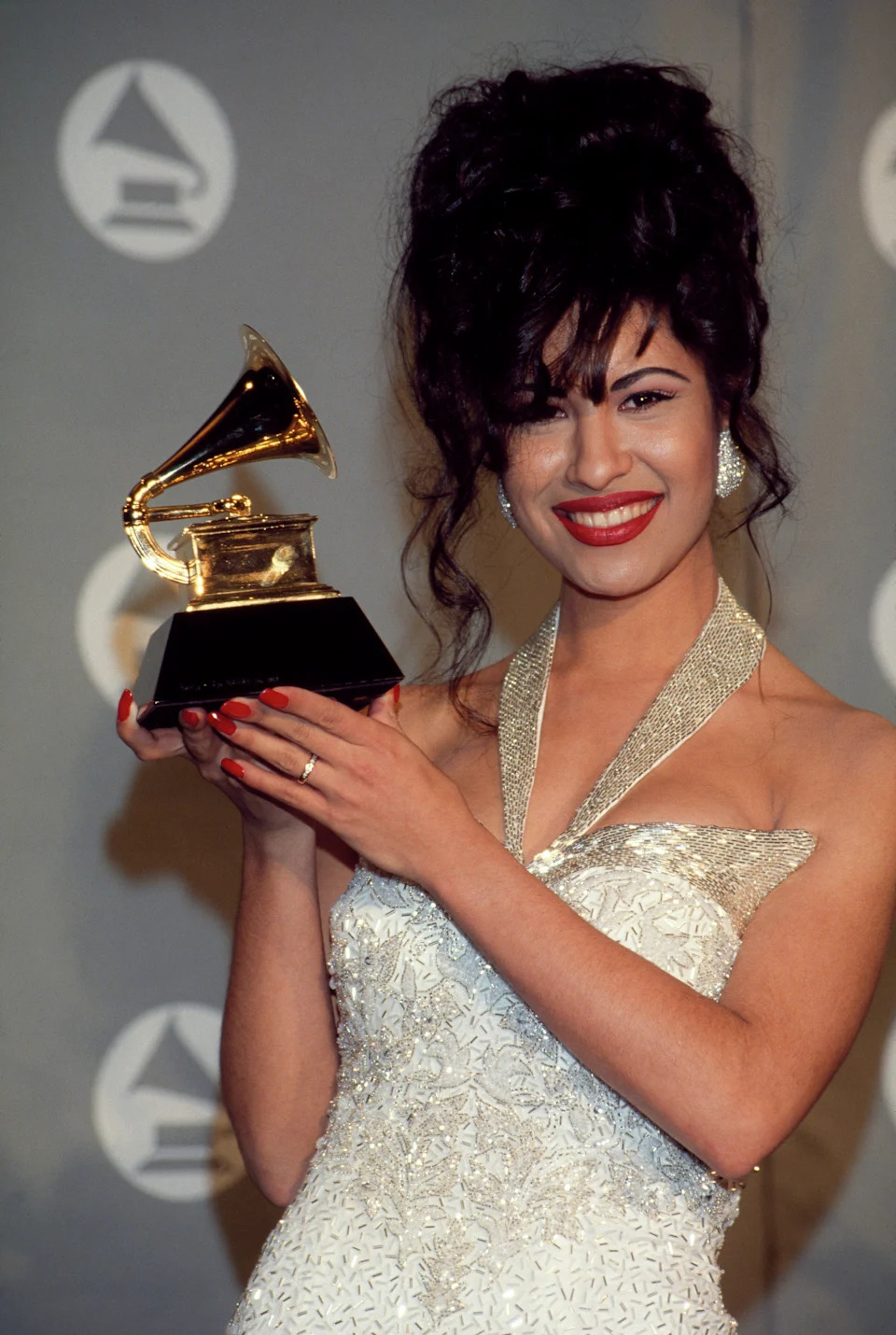 Selena in an elegant gown holds a Grammy award, smiling at a press event