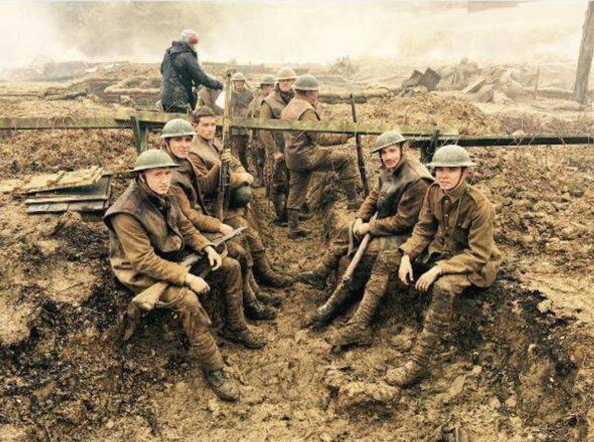 A group of individuals in military attire, possibly soldiers, are seated on the ground in a muddy environment, likely in a trench. One individual stands behind them, possibly observing or assisting. The setting appears to be a war zone or a training exercise, with a blurred background.