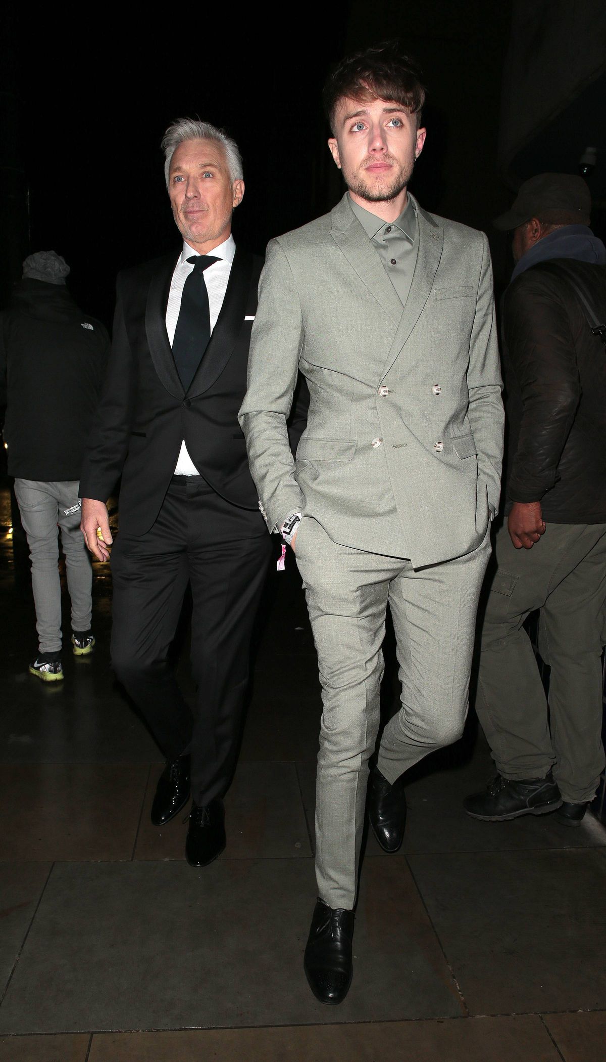 Two individuals dressed in formal attire are standing together in a dimly lit environment. One individual is dressed in a black suit with a tie, while the other is wearing a gray suit. They are facing the camera with several other people in the background, partially obscured by the darkness.