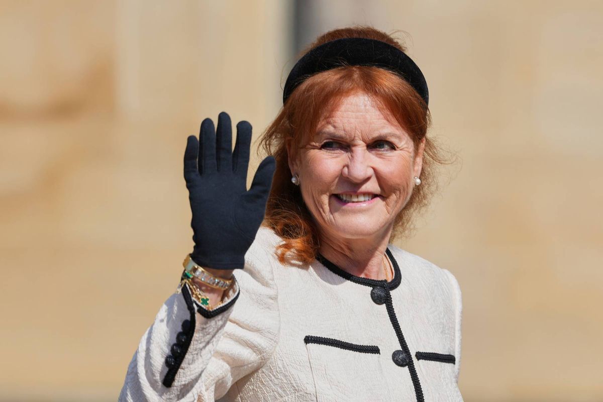 An individual with red hair, donning a white coat with black trim and a black headband, is seen smiling and waving while wearing a black glove.