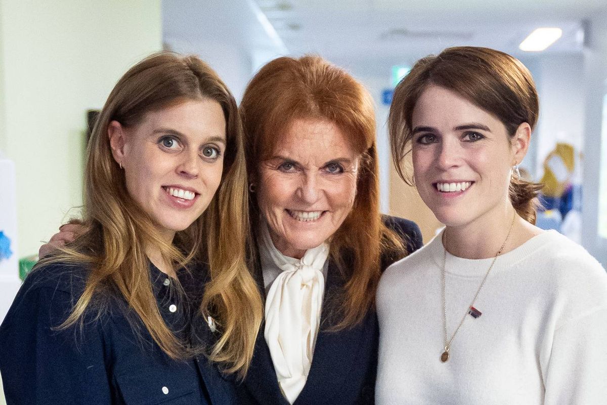 Three women standing together, all smiling and posing for a photograph. The woman on the left has long blonde hair and is wearing a blue jacket, the central figure has red hair and is dressed in a black suit, and the woman on the right has brown hair and is wearing a white top and necklace.