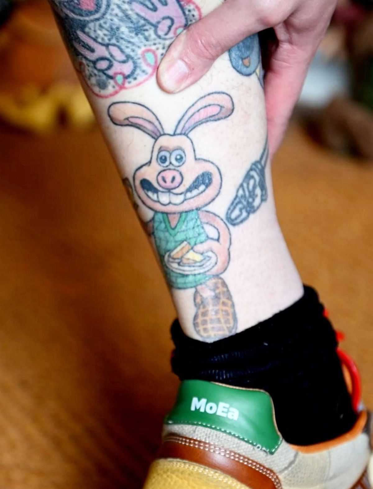A person's lower leg with a detailed tattoo of a rabbit character is partially covered by a black sock. The tattoo features vibrant colors and intricate designs. The individual is wearing a sneaker with a green accent and the word "HOKA" on the side.
