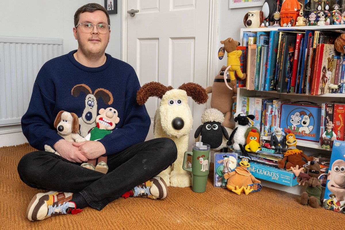 An individual dressed in a blue long-sleeve shirt is seated on the wooden floor, surrounded by a variety of stuffed animals and toys. A bookshelf filled with books and other items is visible in the background.