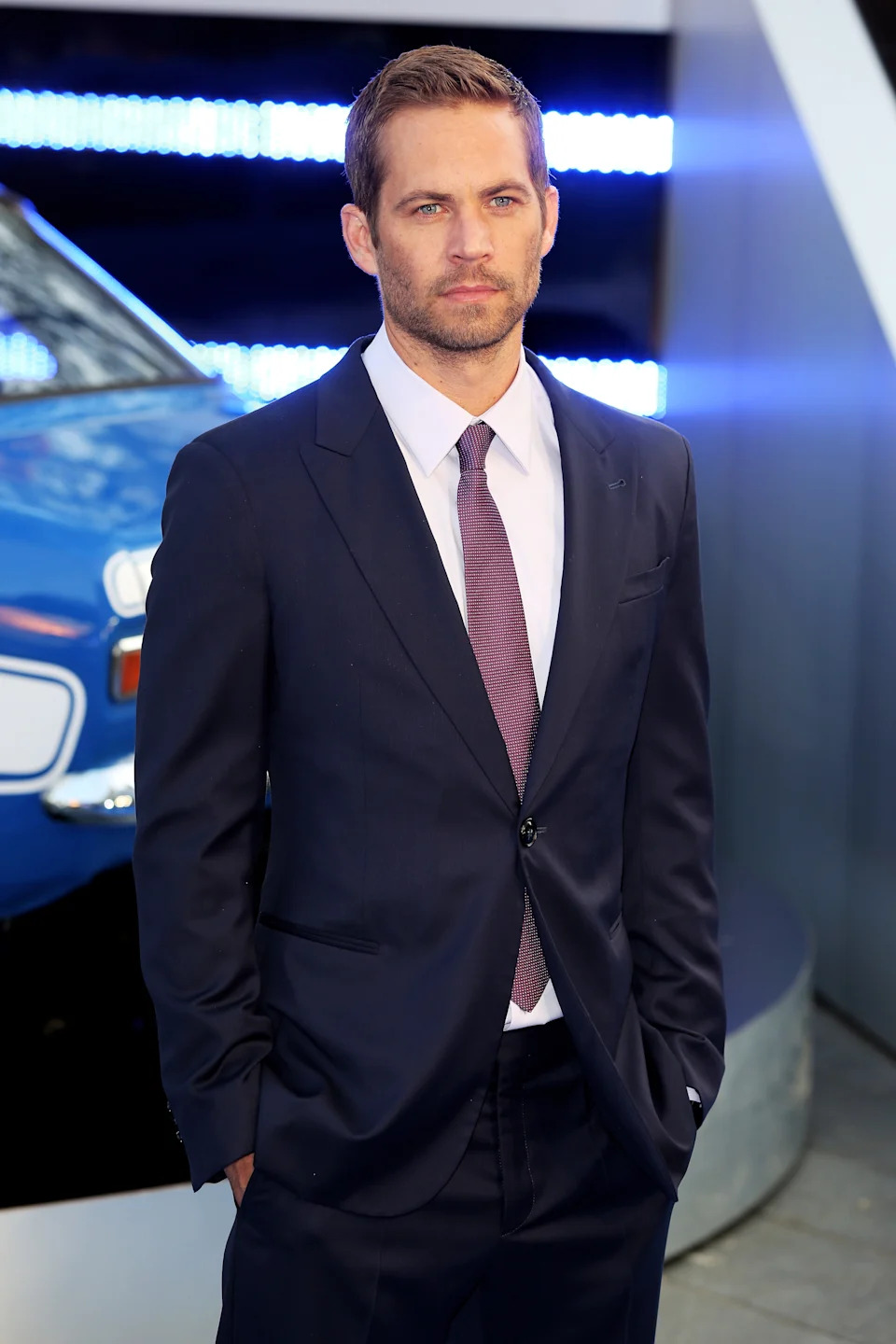 A man in a suit with a tie stands on a red carpet, with hands in his pockets, against a backdrop of a car and bright lights