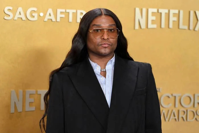 Person in a tailored black suit with long hair and sunglasses on a press event red carpet with industry logos in the background