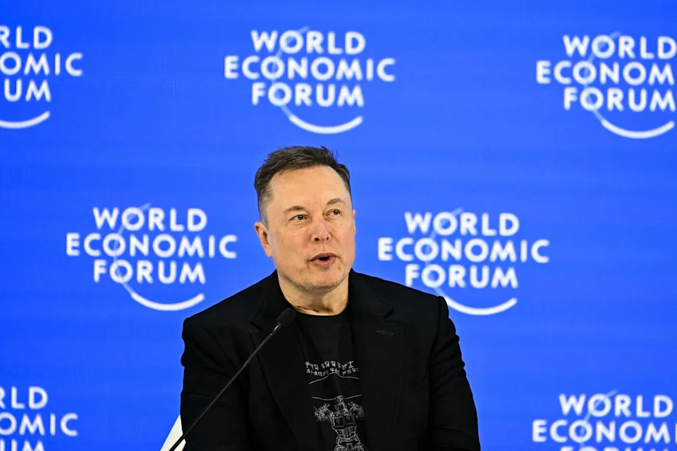 Elon Musk speaking at World Economic Forum, seated at a podium, wearing a dark suit and graphic shirt