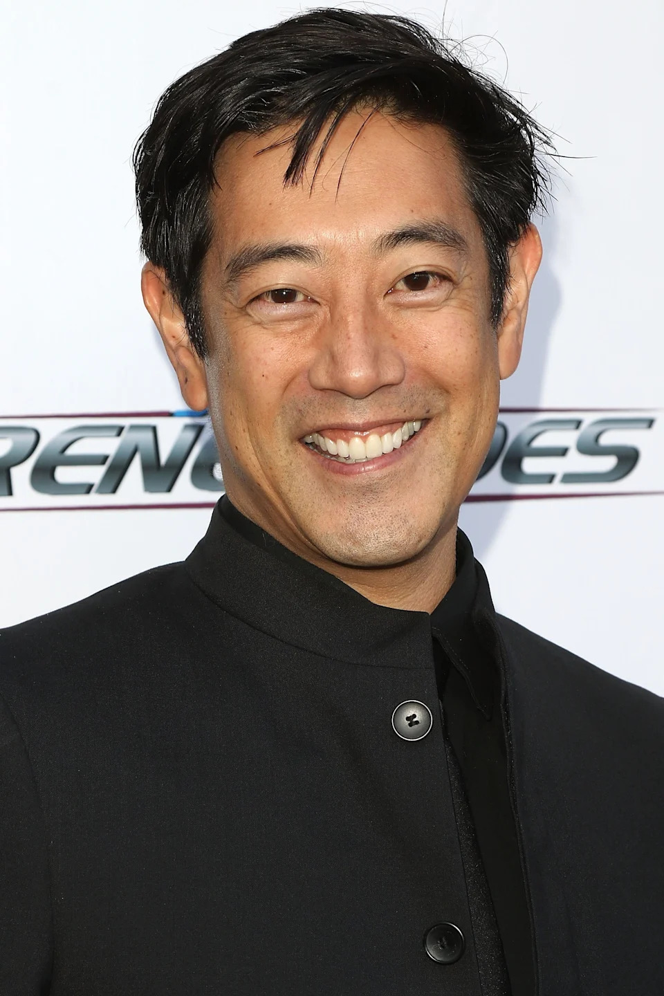 Grant Imahara smiling in a collared outfit at a media event backdrop
