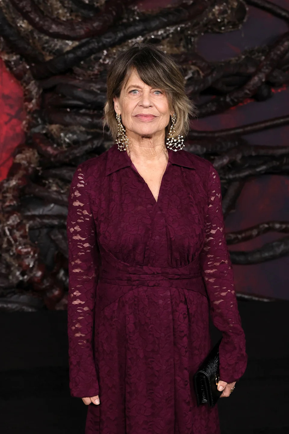 Person in a long lace dress with intricate earrings on a red carpet. They hold a clutch and stand in front of a textured backdrop