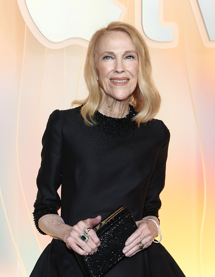 Catherine O’Hara in a black dress holding a clutch, smiling at an event with a light-colored background. Catherine O’Hara in a black dress holding a clutch, smiling at an event with a light-colored background.