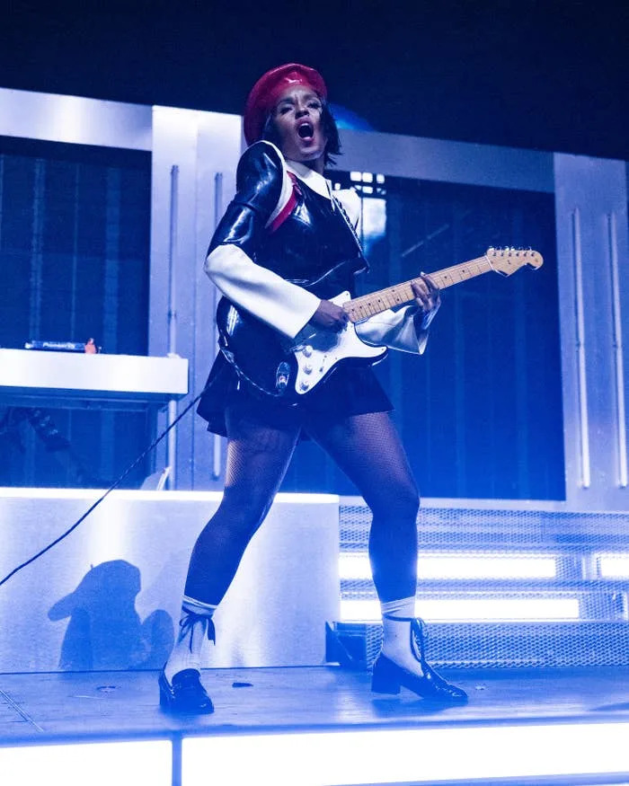 Janelle Monáe on stage wearing edgy, futuristic outfit with short skirt and beret, energetically playing electric guitar
