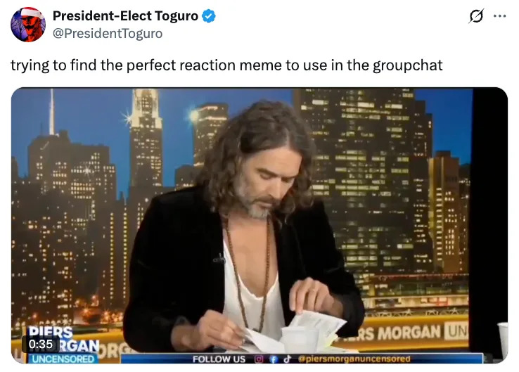 Man with long hair and a blazer sitting at a desk looking through papers, with a cityscape backdrop. Tweet above says, "trying to find the perfect reaction meme."