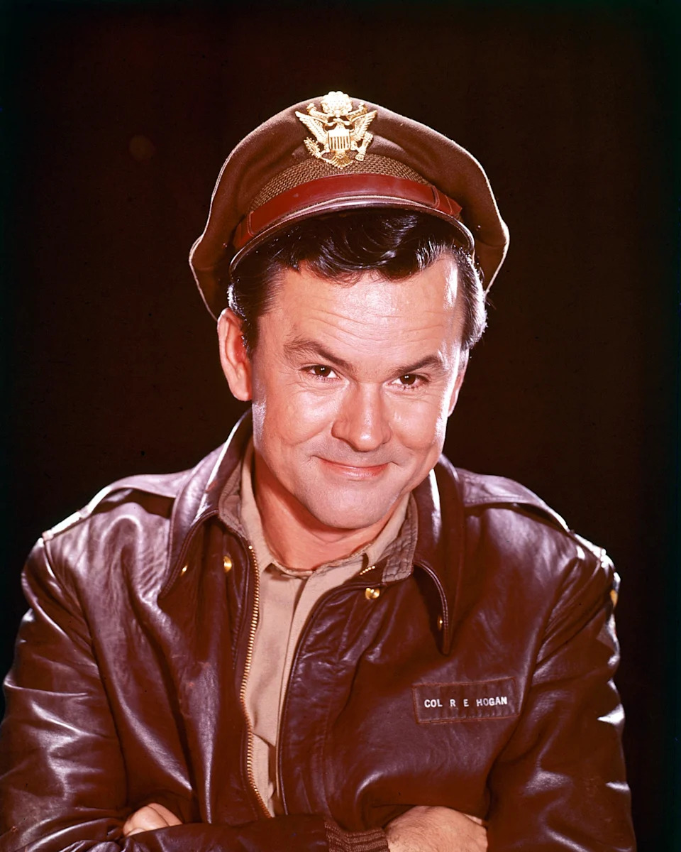 Bob Crane wearing a military-style leather jacket and cap, smiling with arms crossed