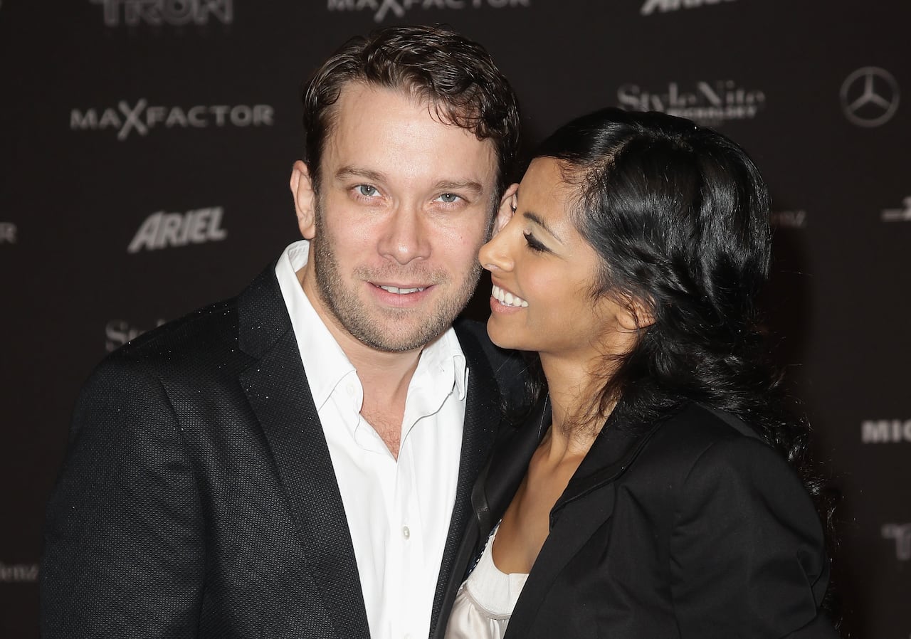 A formally dressed couple stands together, with the woman looking at her husband smiling while he stares into the camera.