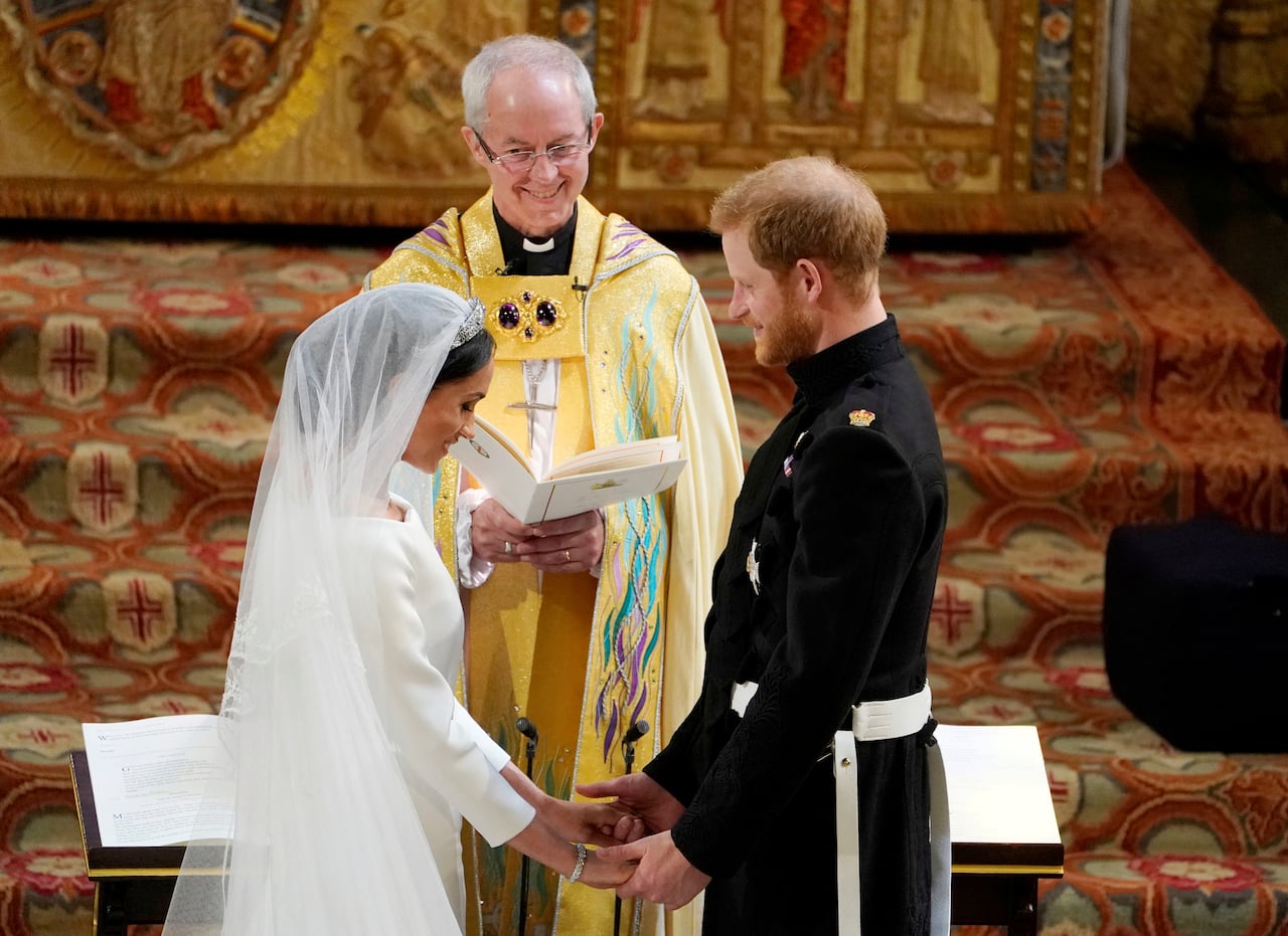 A bride and groom hold hands in front of an altar as a person in religious vestments holding an order of service looks at them.