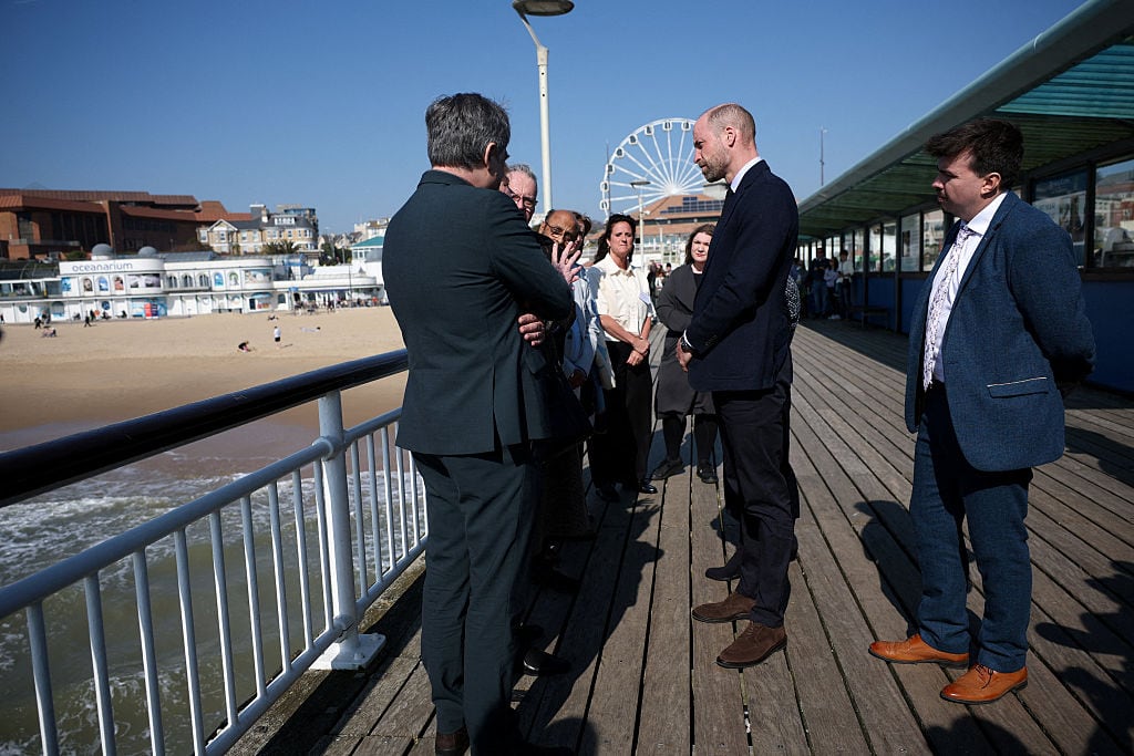 People stand and talk on a pier with a ferris wheel in the background.