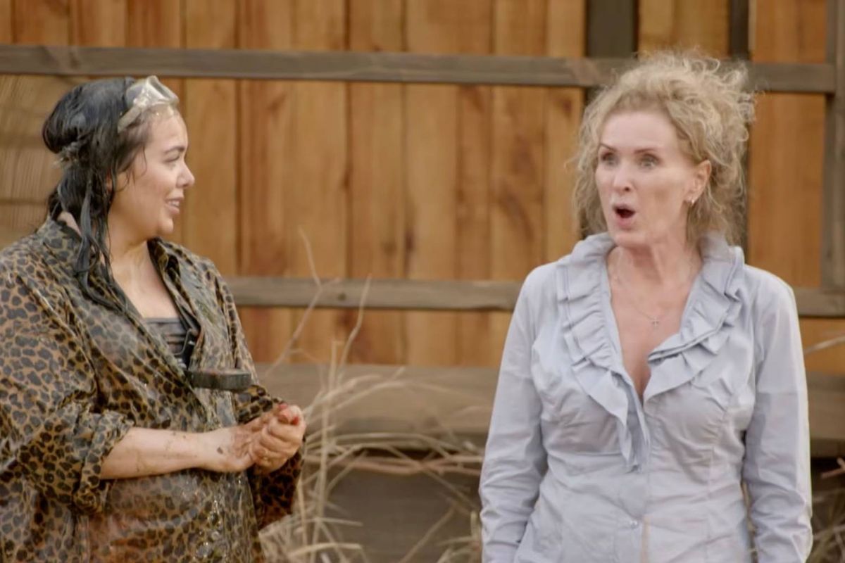 An adult female with curly blonde hair appears to be speaking or shouting with her mouth open, standing in front of a wooden wall.