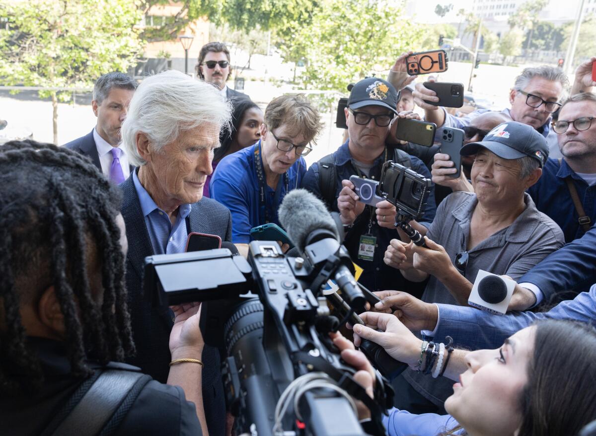 A man with white hair talks in front of a collection of microphones.