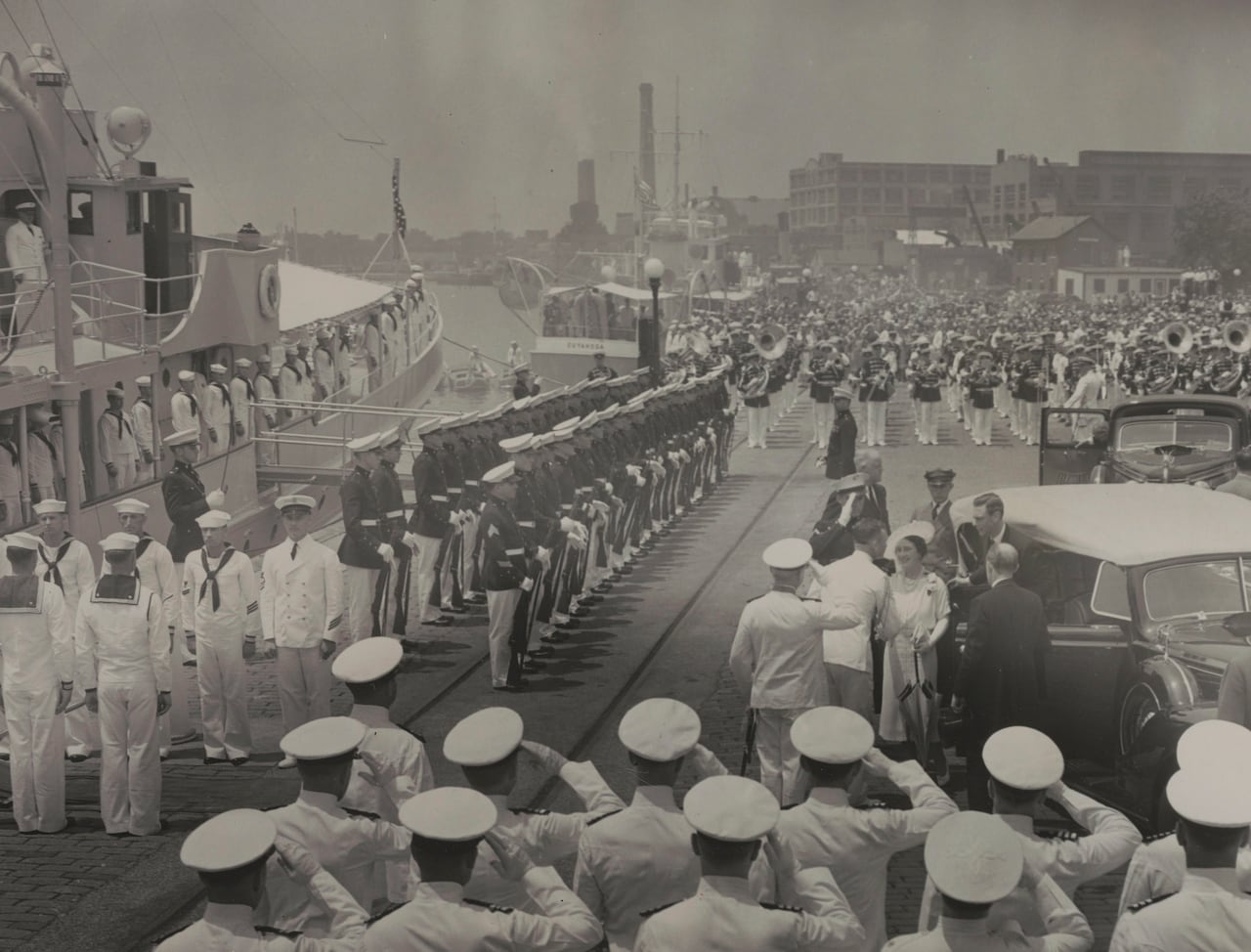 A large group of sailors salute as people are greeted after getting out of a car.