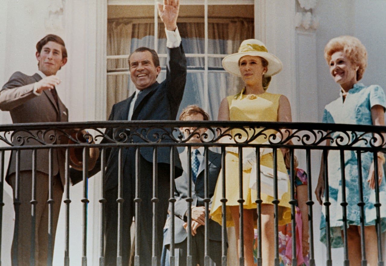 One person waves as they stand with three other people on a balcony.