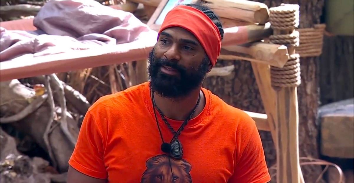 An individual, dressed in an orange t-shirt and a red headband, stands in front of a rustic wooden structure, possibly engaged in conversation or observation. The backdrop features additional wooden elements and what appears to be a makeshift shelter or dwelling.
