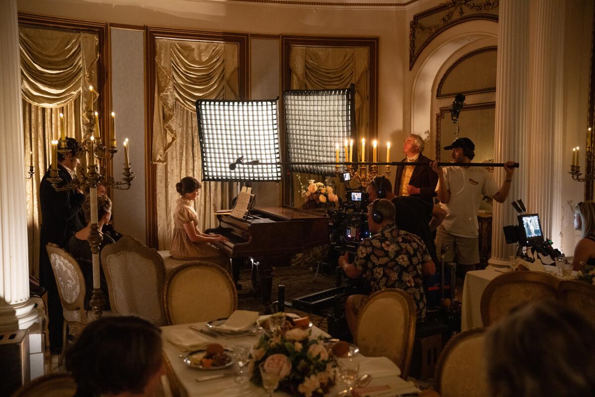 A woman plays at a piano as a long boom mic and lights stand near her. Several camera operators film her.